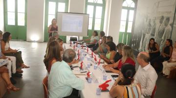 secretários sentados em  mesa olhando apresentação #paratodosverem