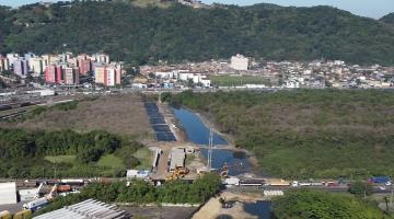 imagem área aparece rio com máquinário de obras ao lado, vasta vegetação ao redor e prédios e morro ao fundo #paratodosverem