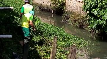 Dois trabalhadores removem resíduos em margem de rio. #Paratodosverem