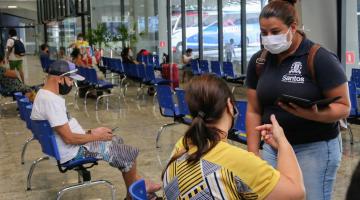 Agente da Ouvidoria conversa com mulher sentada na sala de espera da rodoviária. Há outra pessoas sentadas no mesmo ambiente. #Paratodosverem