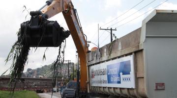 Escavadeira cheia de detritos está elevada em movimento para depositá-los no caminhão que está à frente. #Paratodosverem