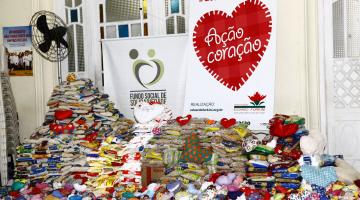 Inúmero pacotes de alimentos empilhados. Ao fundo, um banner da Ação do Coração e outro, do  Fundo Social de Solidariedade. #Pracegover