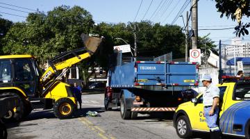 Máquina retroescavadeira transfere materiais recolhidos para caminhão. Acompanhando o trabalho está o carro e um agente da CET. #Pracegover