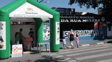 movimento de rua com mãe passeando com dois filhos, à esquerda uma tenda verde e branca