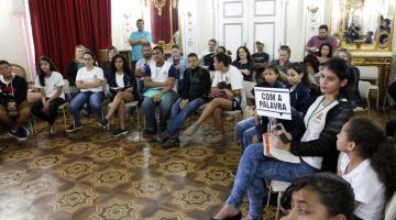 Alunos em círculo durante mediação. Uma delas segura placa dizendo com a palavra #pracegover 