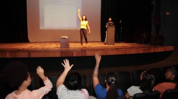 em um palco mulher com mão levantada, na platéia , de costas mulheres sentadas e com mãos levantadas