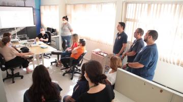 pessoas sentadas em uma sala , mesa ao fundo, à direita três pessoas em pé #paratodosverem