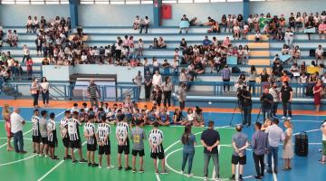 jogadores na quadra e diversos estudantes os olhando na arquibancada #paratodosverem