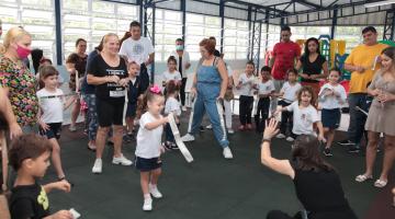 criança uniformizada brincando com adulto ao redor de vários adultos em uma quadra #paratodosverem