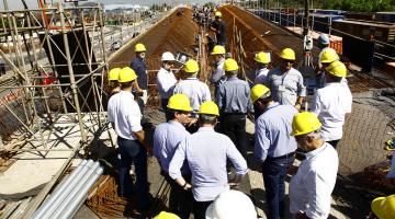 Homens e mulheres usando capacetes estão diante da obra de uma ponte. Há uma grande estrutura de ferros. #Pracegover