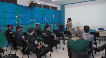 sala de aula com guardas municipais uniformizados sentados, e outras pessoas. Ao fundo, uma mulher está em pé falando ao grupo. #paratodosverem