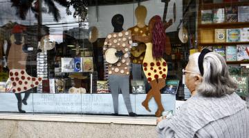 Figuras em papelão e coloridas expostas em vitrine. Uma mulher está olhando do lado de fora. #Pracegover