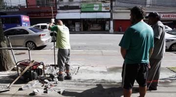 Homem está cavando buraco em trecho da calçada para instalação de barra. #Pracegover