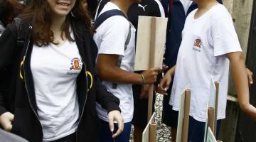 Alunos uniformizados estão em rua diante de placas informativas. #Pracegover