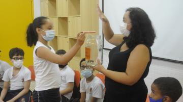criança coloca papel em pote de vidro nas mãos da professora #paratodosverem