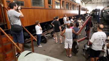 Visitantes na garagem de bonde ouvem explicações de guia de turismo e observam vagões antigos. #Paratodosverem