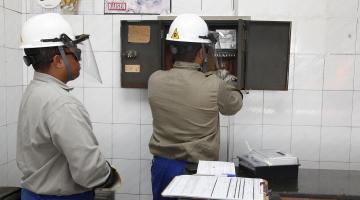 Funcionários da CPFL, com capacete e protetor visual verificam relógio de luz em estabelecimento. #Paratodosverem