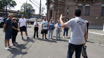 guia usando camiseta da seectur está falando a grupo de pessoas em frente à Bolsa Oficial do Café. #paratodosverem 