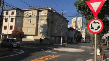 Placa de minirrotatória em primeiro plano, com dispositivo ao fundo durante passagem de veículo. #pracegover