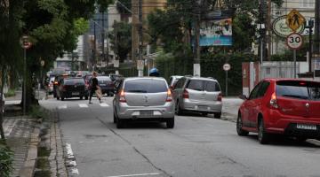 Rua com carros estacionado à direita e outros circulando. #paratodosverem