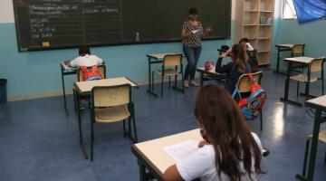 sala de aula com crianças sentadas, todas de costas. A professora está ao fundo, à frente da lousa e falando aos alunos. #paratodosverem