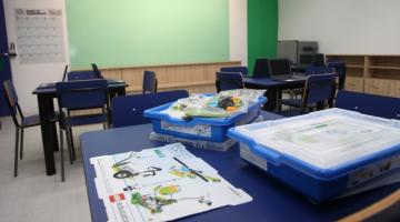 Sala de aula com mesa em primeiro plano. Sobre ela há duas caixas e um desenho. Ao fundo, a lousa. #Paratodosverem
