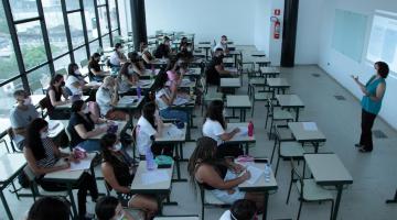 sala de aulas com alunos sentados e professora à frente. Sala é fechada por vidro que permite visualização de parte do centro histórico. #paratodosverem