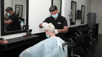 homem está em pé vestindo o uniforme preto do salão. Ele corta o cabelo de um rapaz que está sentado. O ambiente tem outras cadeiras para corte de cabelo. #paratodosverem