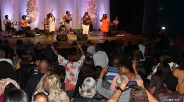 na plateia, público acompanha apresentação em pé, com mãos para o alto e alguns filmando com celular. #paratodosverem 