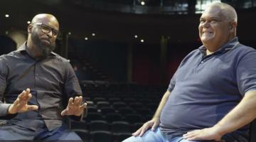 Cantor conversa com o entrevistador em um palco. #paratodosverem