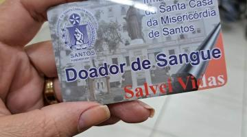 mão segura cartão de doador onde se lê Doador de Sangue. Salve vidas. Há um símbola da Santa de casa de santos no alto. #paratodosverem 