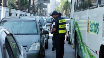 guardas municipais estão em pista de avenida, com carros parados. Eles estão entre carros e ônibus distribuindo folhetos. #paratodosverem