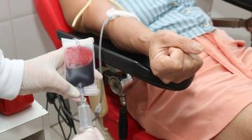 close em doação de sangue. Mãos com luvas seguram bolsa cheia de sangue. Uma mulher está sentada a uma cadeira, com o braço estendido no suporte. Um duto da bolsa está ligado ao seu braço. #paratodosverem