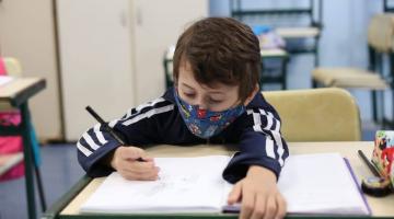 Criança de máscara escreve em um caderno em uma sala de aula #paratodosverem