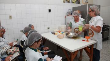 Duas cozinheiras preparam pão enquanto alunas observam sentadas #paratodosverem