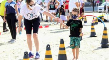 muilher segura mão de criança com deficiência visual, que é conduzida para caminhar entre cones na areia da praia. #paratodosverem