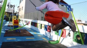 menina está brincando em balancê. Seus pés e corpo estão no ar, em primeiro plano e no alto. Ao fundo, os novos brinquedos. #paratodosverem