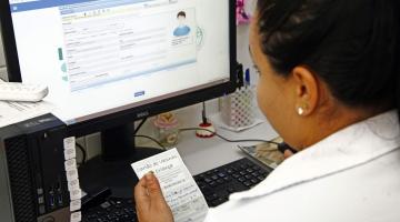 mulher segura carteira de vacinação diante de computador onde ela preenche dados. #paratodosverem