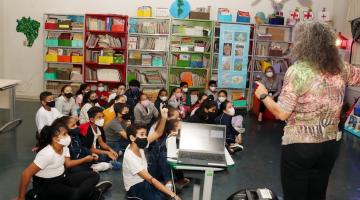 ALUNOS ESTÃO SENTADOS NO CHÃO ATENTOS A UMA MULHER QUE FALA EM PÉ. uM DELES LEVANTA A MÃO PARA PARTICIPAR. A sala tem prateleiras com livros e decoração voltada para o meio ambiente. #paratodosverem