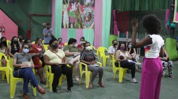 Mulheres sentadas observam instrutora em palestra na quadra da escola de samba União Imperial. #pratodosverem