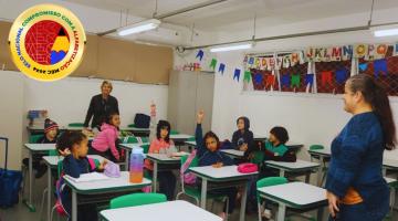 sala de aula com alunos e professores à frente. Selo está sobreposto sobre a imagem, no alto, à esquerda. #paratodosverem 