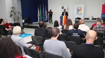Palestrante em púlpito coberto com bandeira com as cores do arco-íris é observado pelo público sentado. #paratodosverem