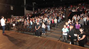 Palestrante no palco do Teatro do Sesc com plateia repleta de educadores. #paratodosverem