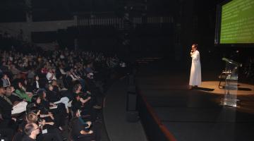 Especialista, no palco do teatro, fala ao microfone para o público na plateia formado por educadores. #paratodosverem