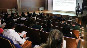 auditório com pessoas sentadas acompanhando palestra. Ao fundo há um telão. #paratodosverem