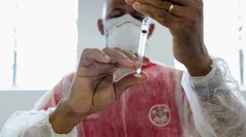 homem insere conteúdo de frasco de vacina em seringa. Ele usa máscara e avental