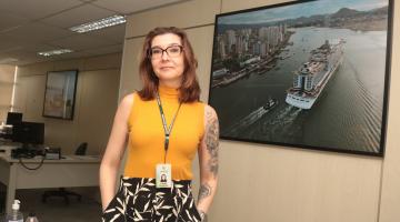 servidora posa ao lado de quadro com navio #paratodosverem