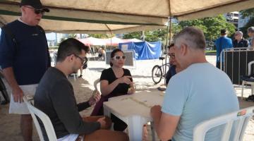 três pessoas estão sentadas à mesa jogando dominó. Um está em pé. Eles estão sob uma barraca na areia da praia. #paratodosverem