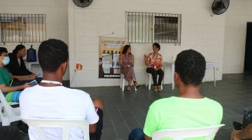 pessoas estão reunidas em um salão. Em primeiro plano, dois rapazes estão de costas para a imagem. Ao fundo, duas mulheres estão sentadas conversando. #paratodosverem