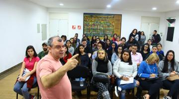 Estudantes estão sentados em auditório e ouvem atentos o palestrante que está à frente de todos. #Pracegover